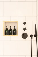 Uma área de duche com três frascos de shampoo e condicionador, um chuveiro manual e um botão de controlo fixado na parede.