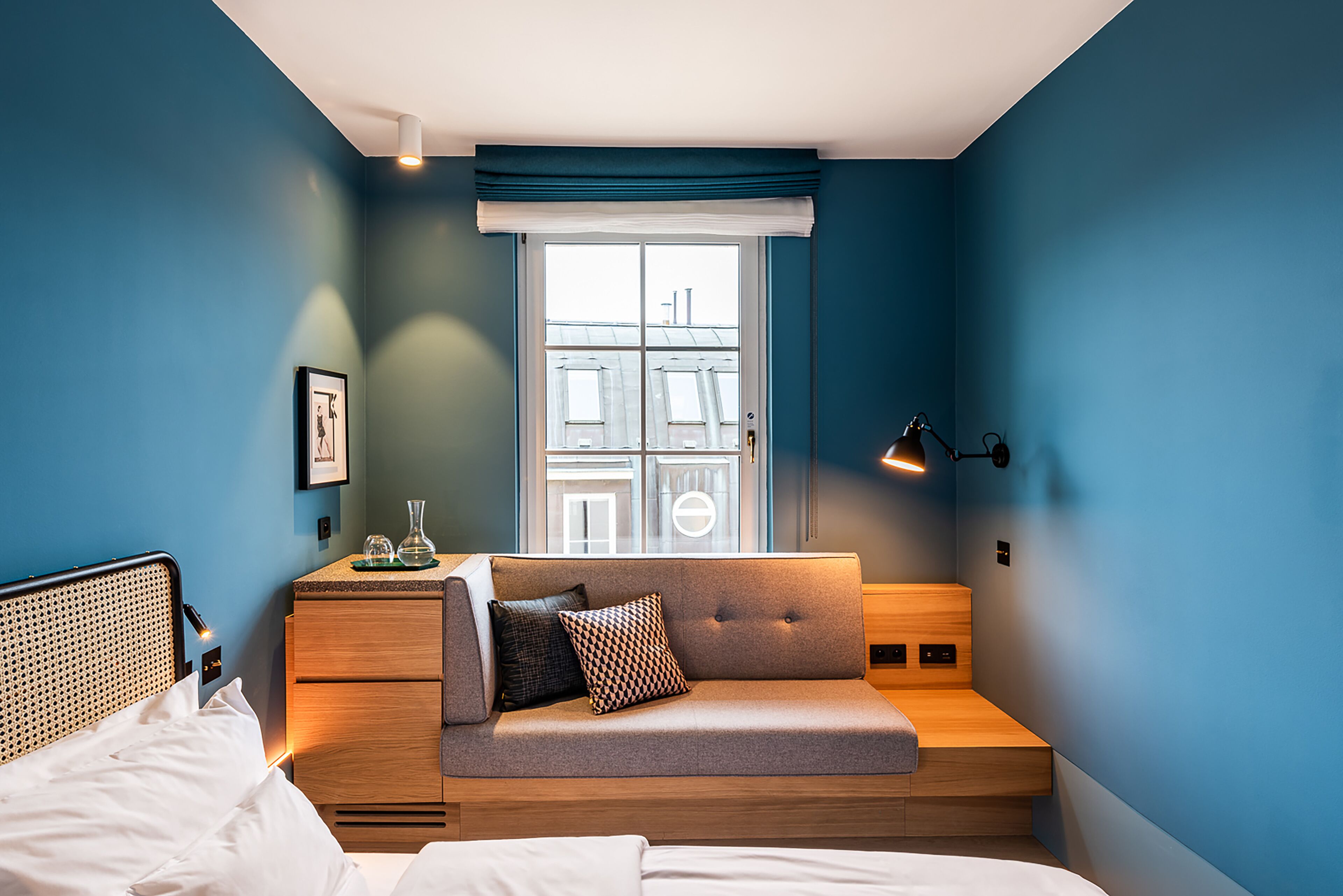 A bedroom with a bed, a wooden nightstand, a sofa bed, a window with blinds, and a wall-mounted lamp.