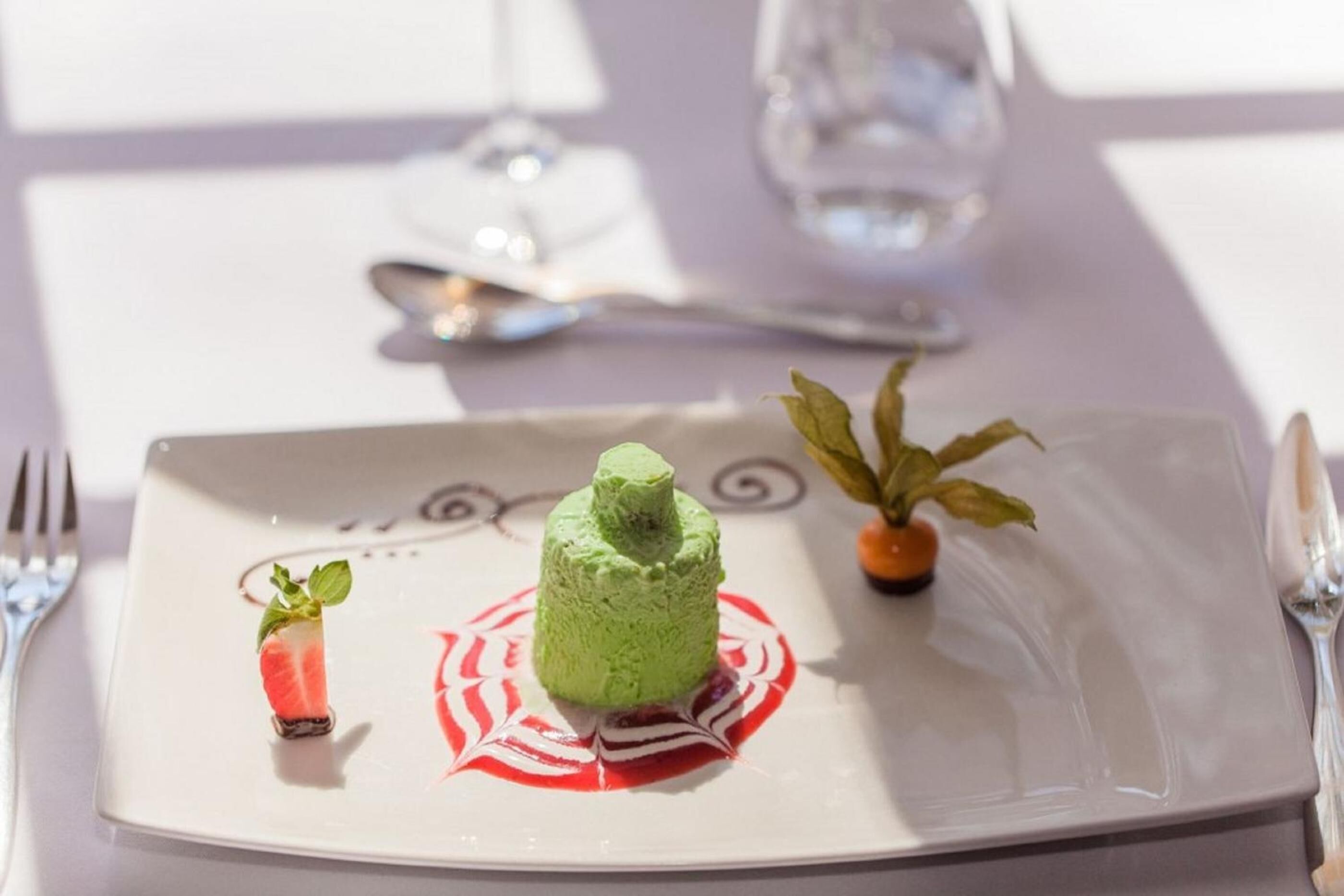 Un postre en un plato blanco con diseño rojo y blanco, una planta pequeña y un vaso de agua sobre una mesa.