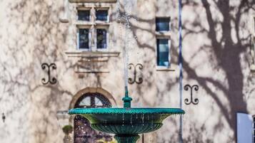 Una fuente con agua brotando desde la parte superior, frente a un edificio con ventanales de arco y detalles decorativos.