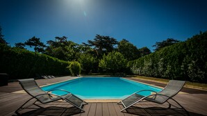 Piscina rectangular con dos sillas reclinables sobre una terraza de madera, rodeada de follaje.