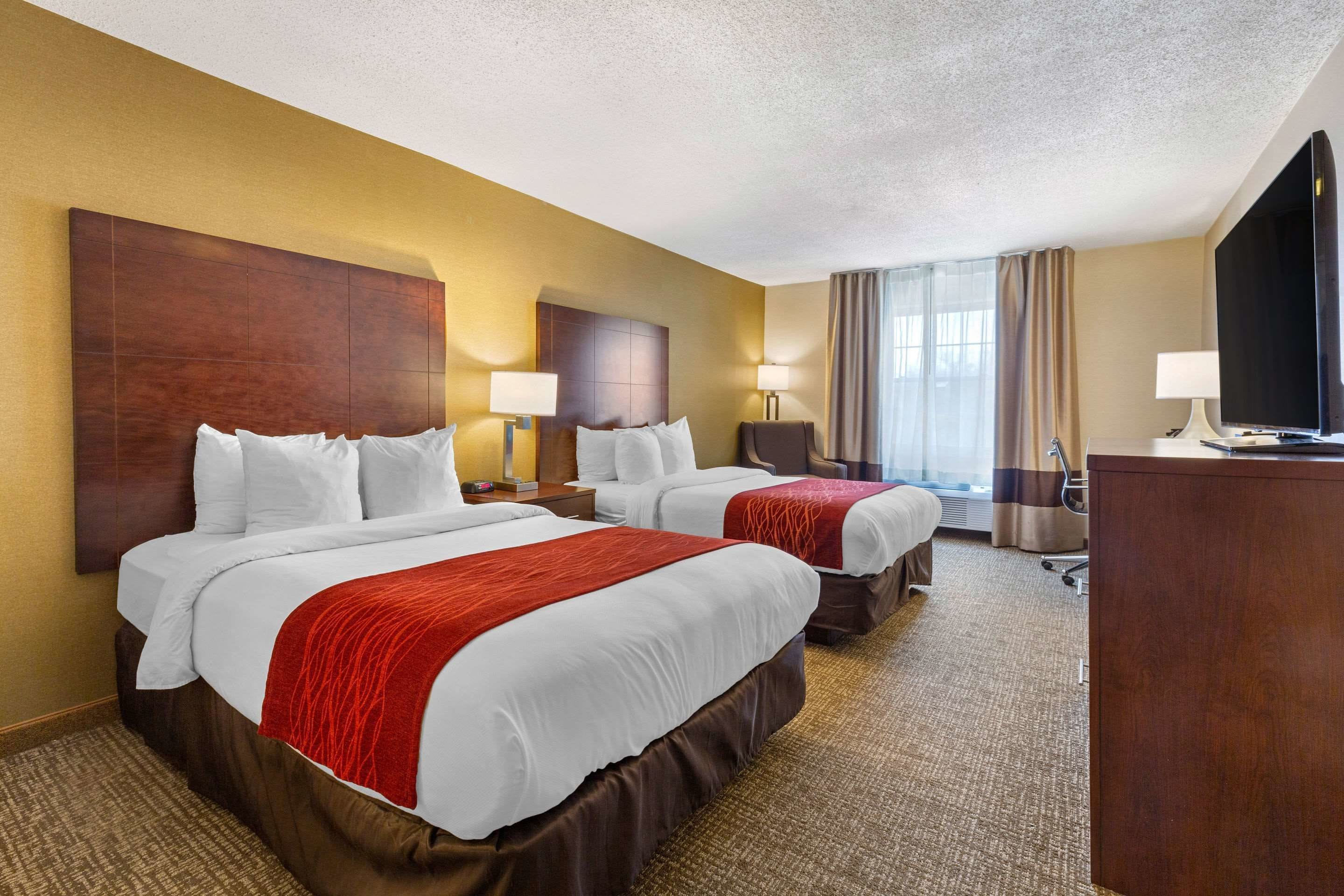 A hotel room with two beds, a desk, a TV, and a large window with curtains.