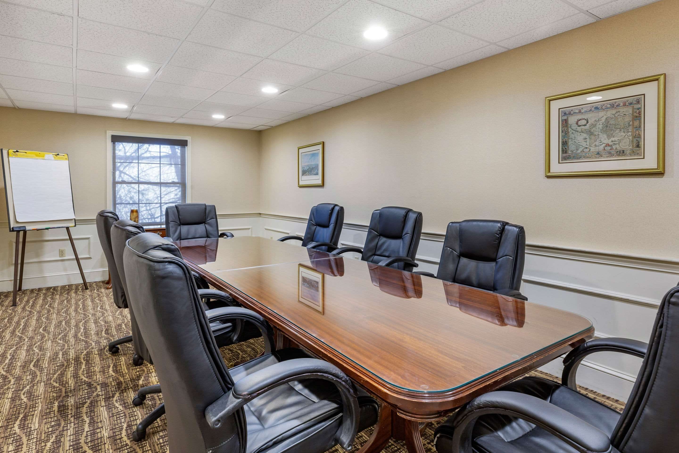 A conference room with a long wooden table, six black leather chairs, a whiteboard, and two framed pictures on the walls.