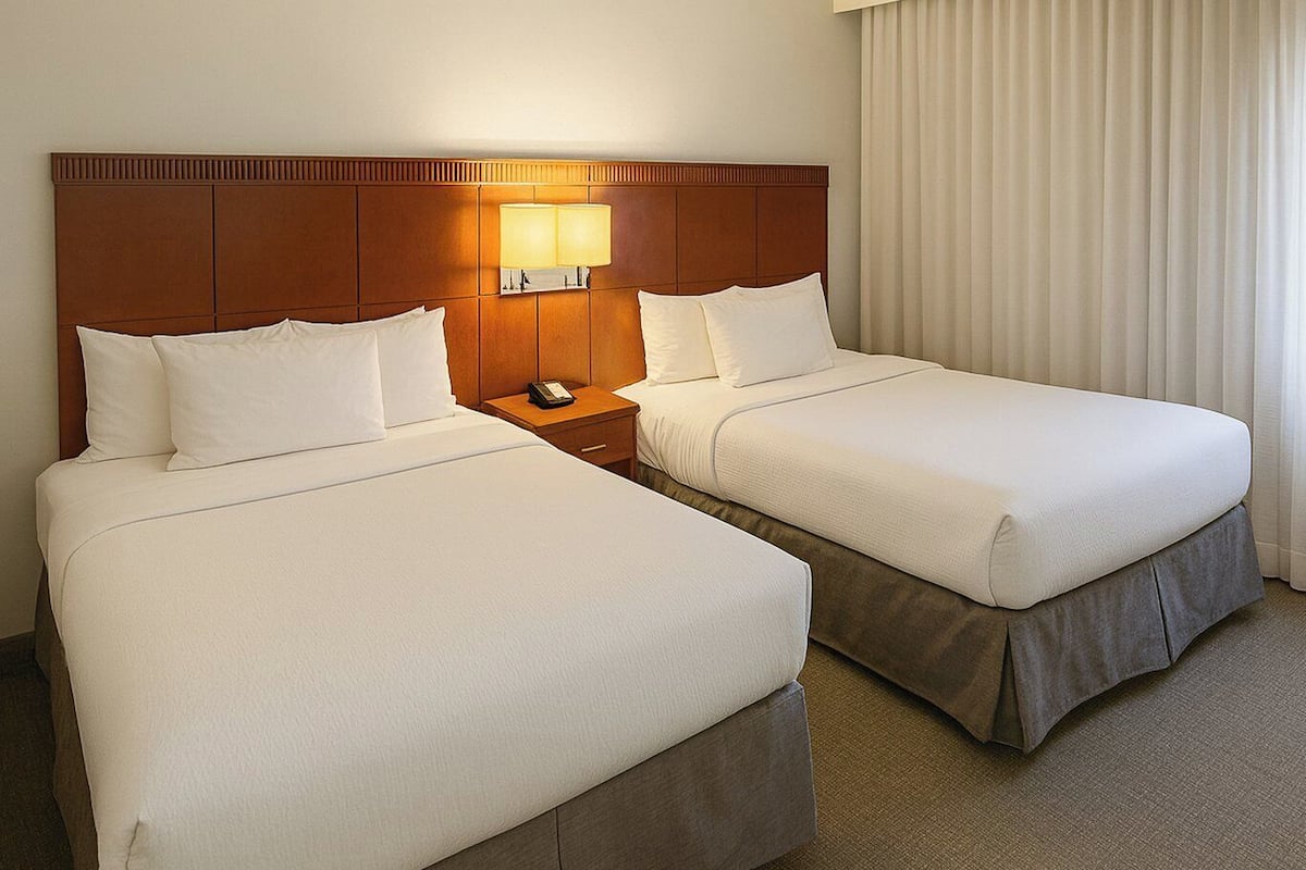 A hotel room with two beds, a nightstand with a lamp, and a window with curtains.