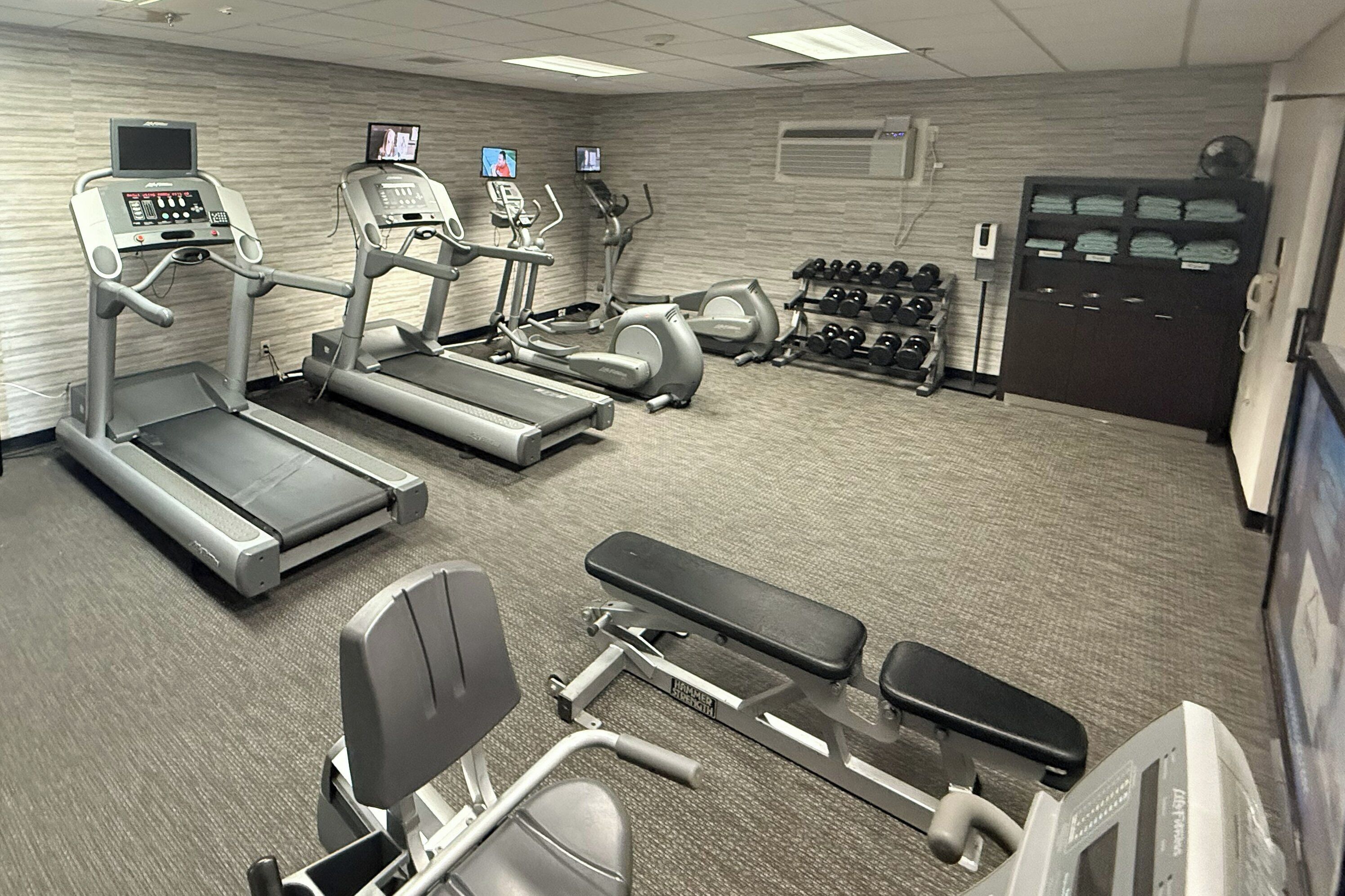 A gym with treadmills, elliptical machines, a bench press, dumbbells, and a TV screen.