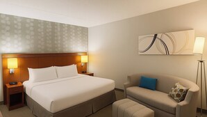A hotel room with a bed, bedside tables, a sofa, an ottoman, a floor lamp, and a wall art piece. - Hotel Preston (Montgomery)