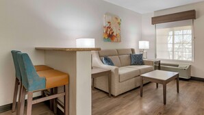 Un salon moderne avec un canapé, une table à manger et une chaise. Il y a une fenêtre donnant sur des arbres et une œuvre d’art accrochée au mur.