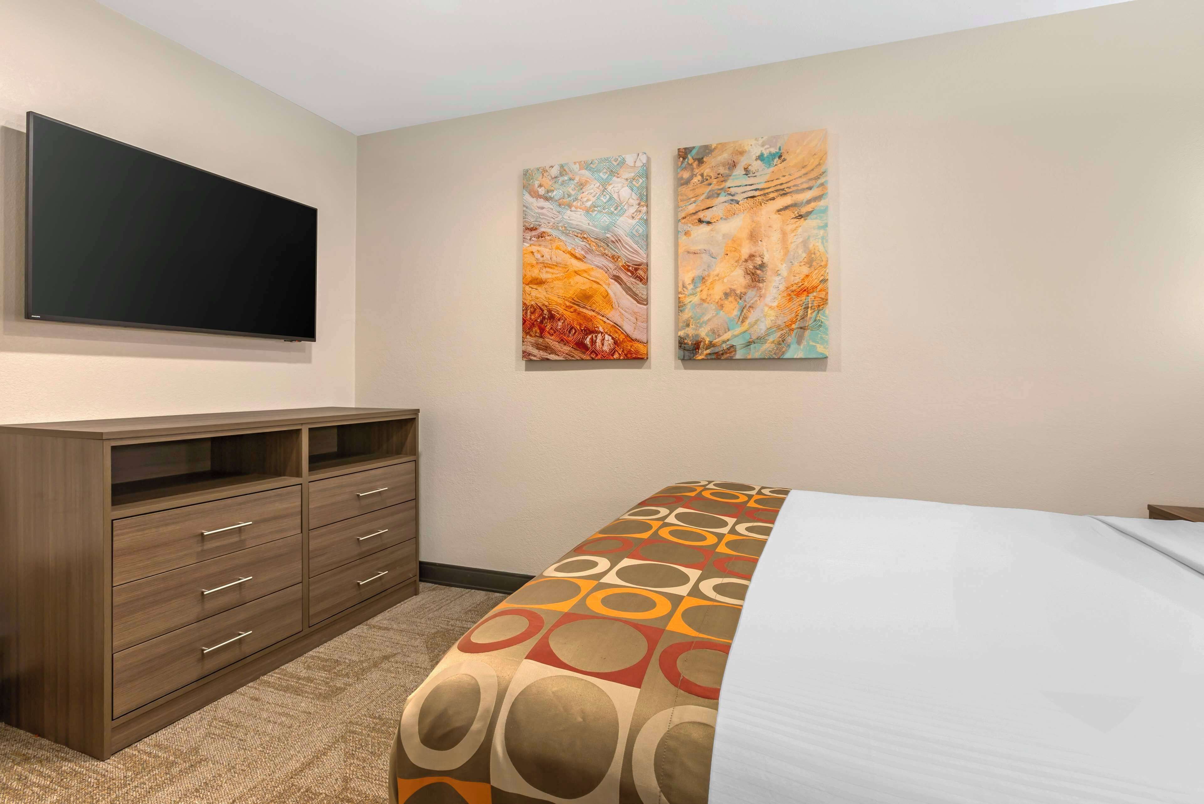 Una habitación de hotel con cama, televisor montado en la pared y dos cuadros abstractos.