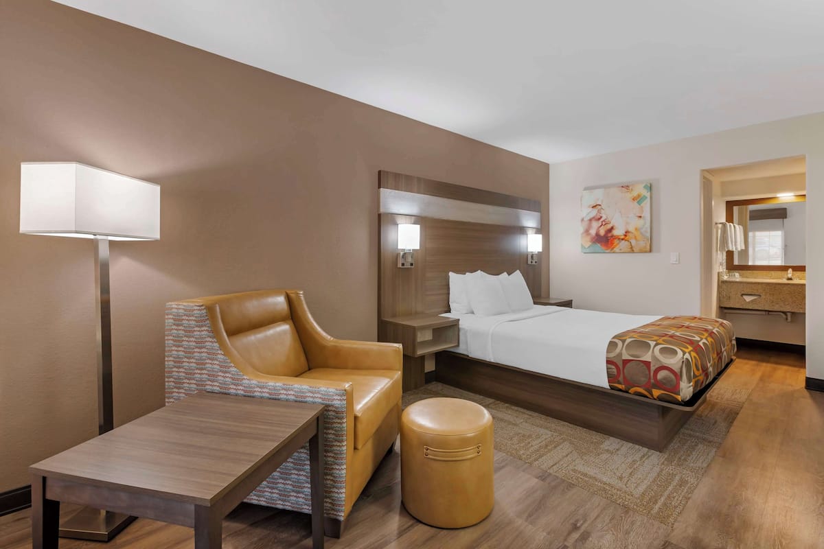A hotel room with a bed, a chair, a table, a lamp, and a small ottoman.