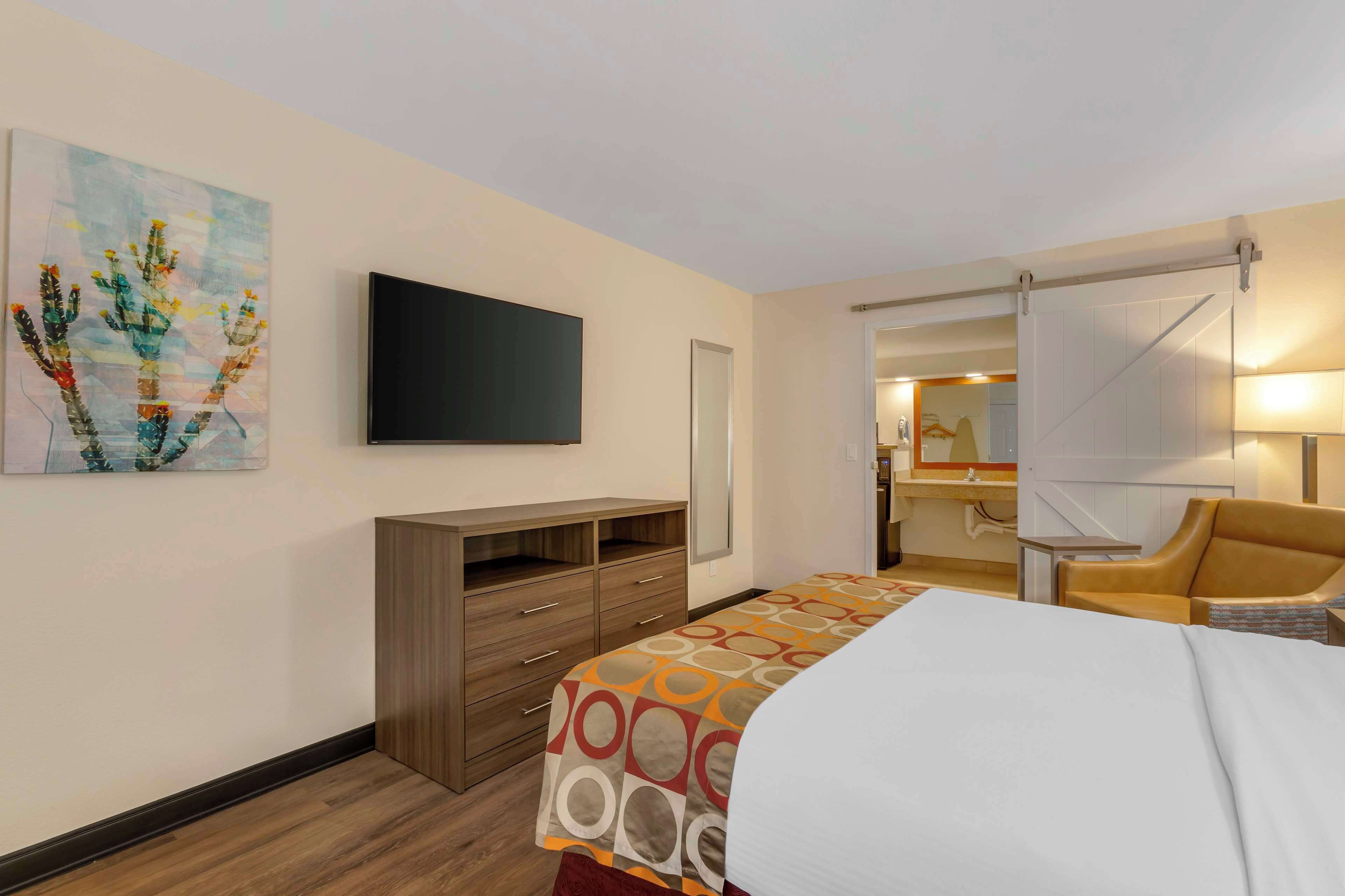 A hotel room with a bed, a TV, a wooden dresser, a chair, and a painting on the wall.