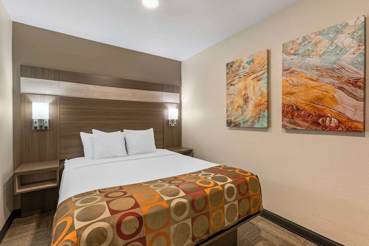 A hotel room with a large bed, a wooden headboard, two wall-mounted lights, and two abstract paintings.