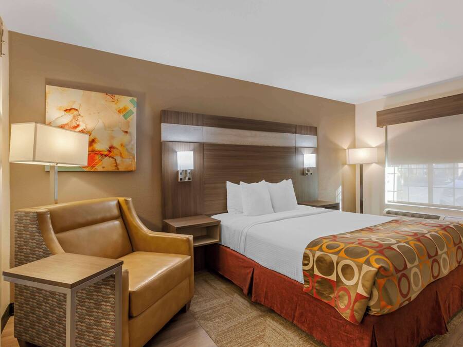 Quarto de hotel com cama, escrivaninha, cadeira, cabeceira e uma obra de arte na parede.