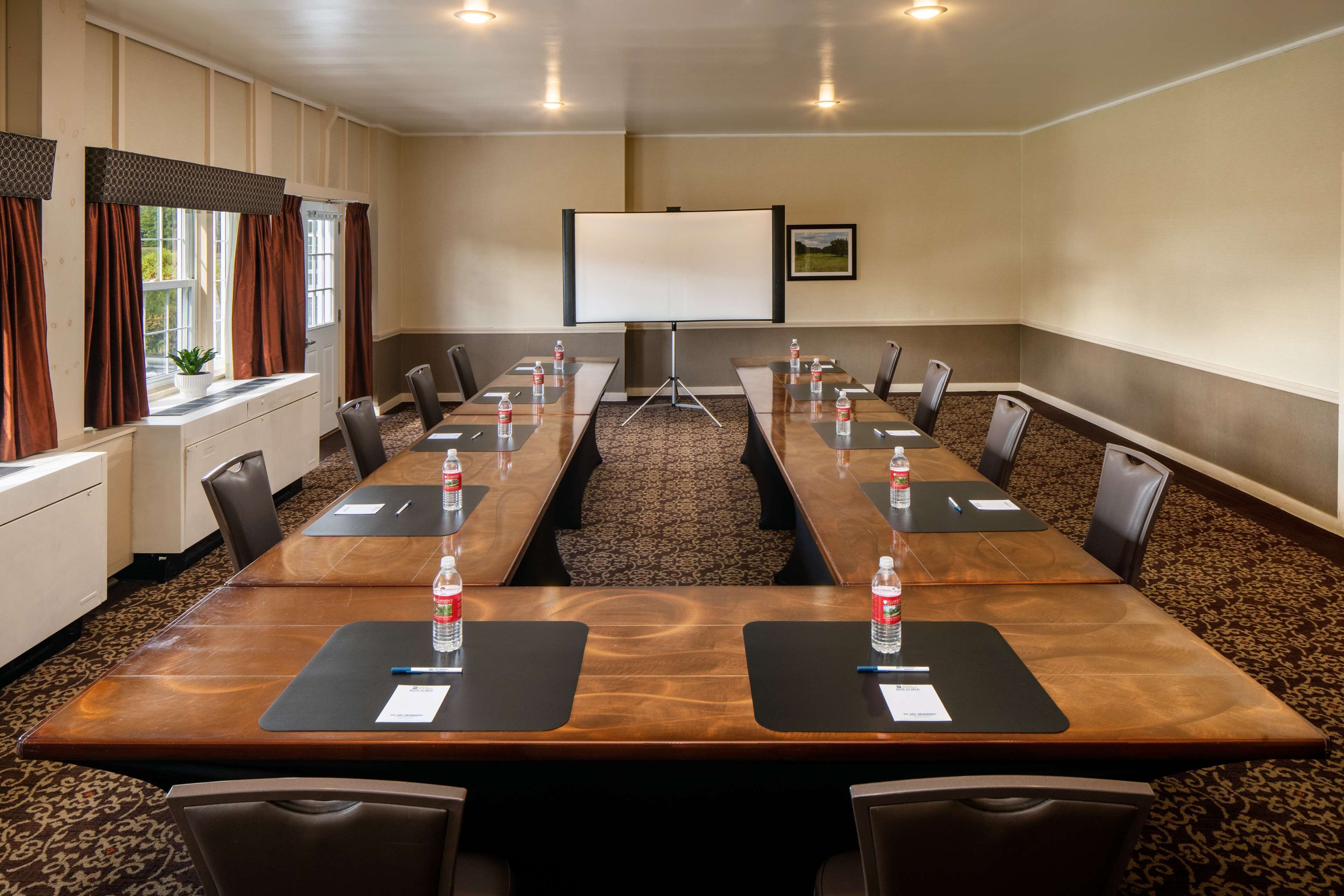 Une salle de conférence avec une longue table, des chaises, un écran de projection et une fenêtre avec des rideaux.