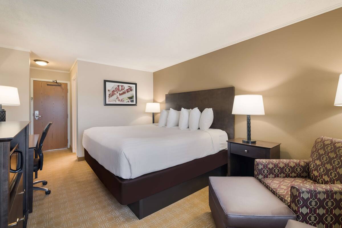 A hotel room with a large bed, two bedside tables with lamps, a desk with a chair, and a patterned armchair.