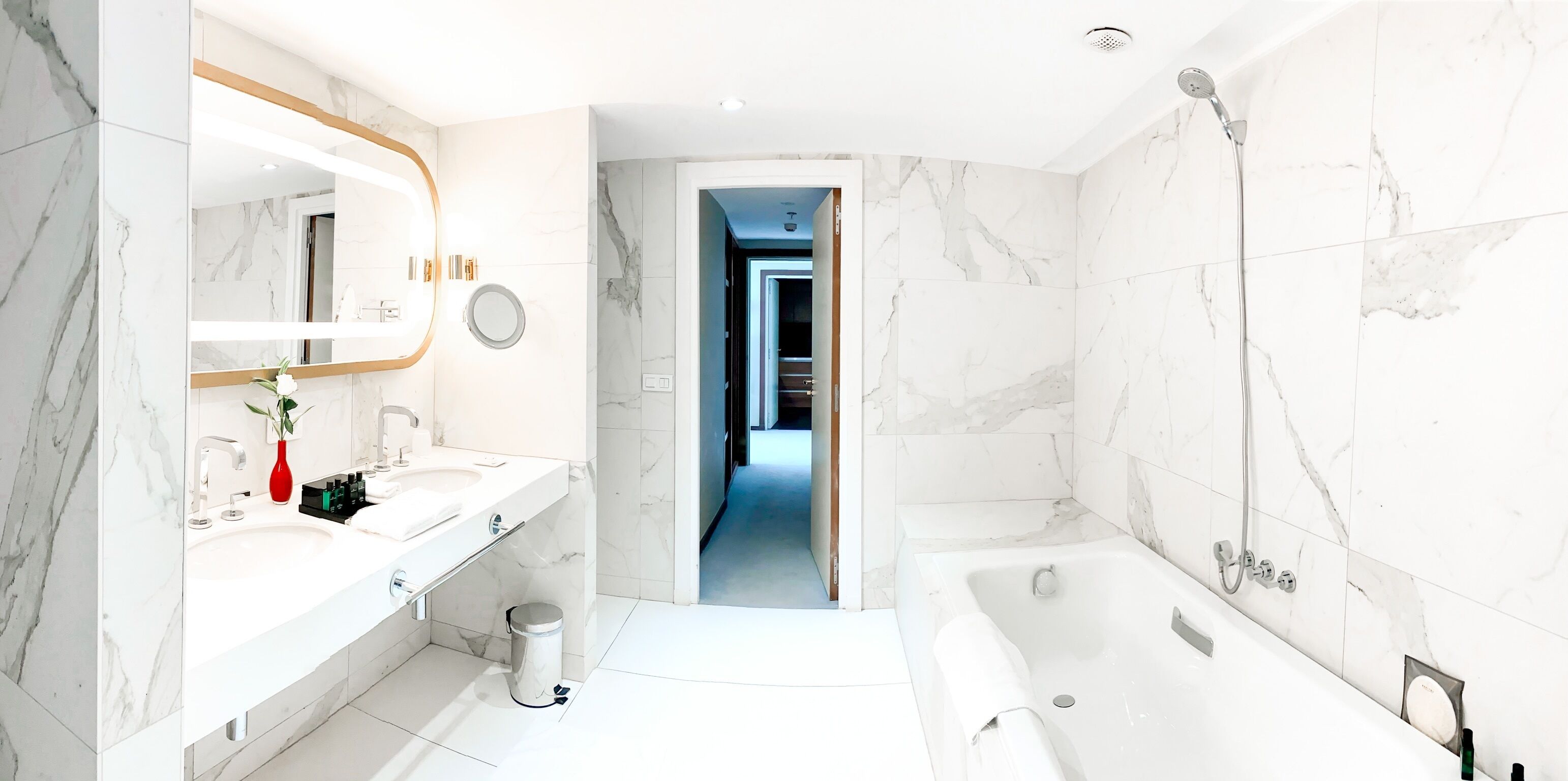 A modern bathroom with a large mirror, white marble tiles, a bathtub, and a vanity with a sink and a red vase.