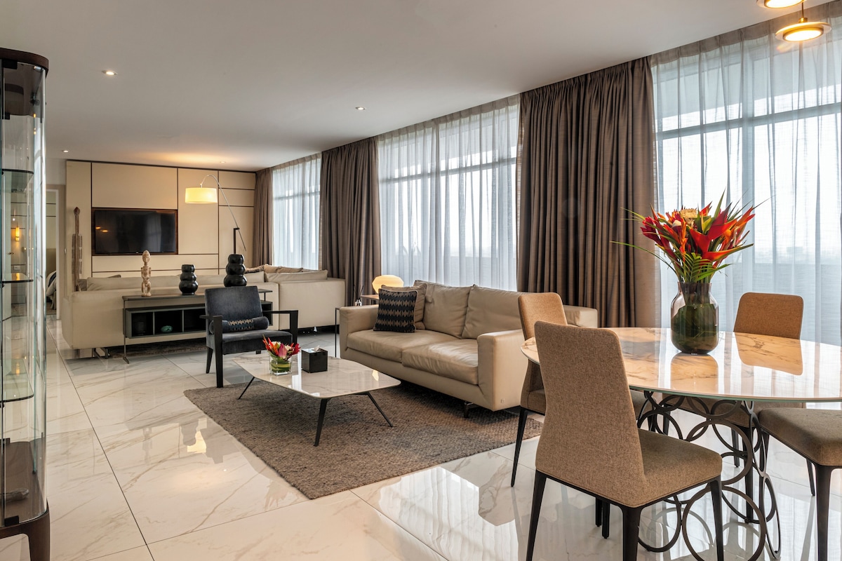 Un salon moderne avec un canapé, une table basse et une table à manger. On y trouve également un téléviseur, un vase avec des fleurs et de grandes fenêtres avec des rideaux.