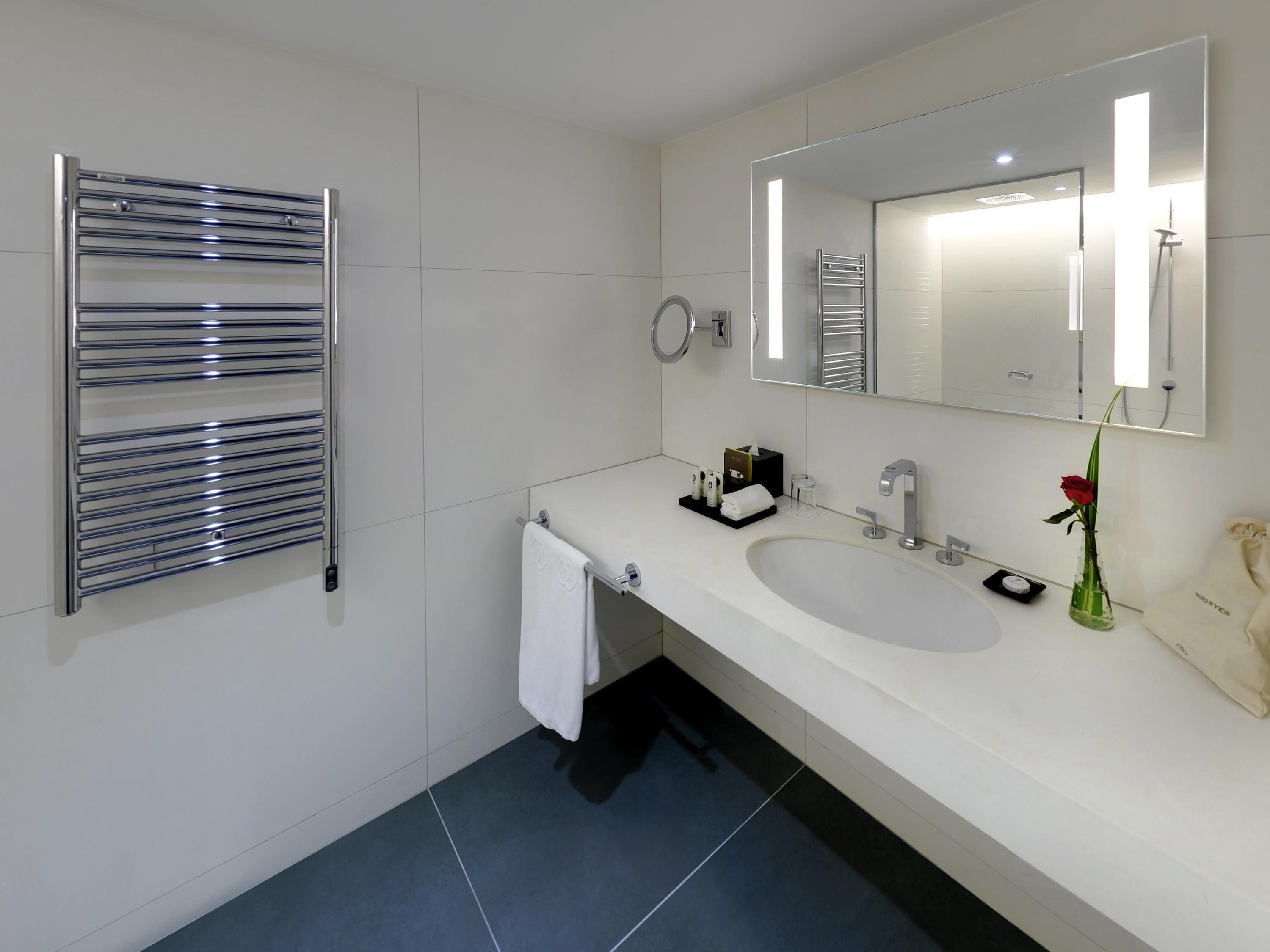 Une salle de bain moderne avec un lavabo blanc, un grand miroir, un porte-serviettes et un vase contenant une seule rose rouge.