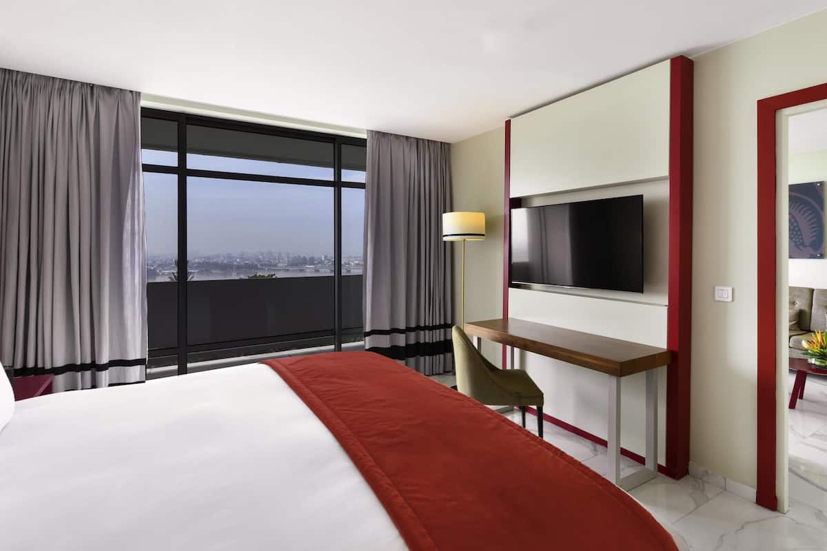 Habitación de hotel con una cama grande, un escritorio, un televisor y vistas a un paisaje urbano a través de un ventanal.