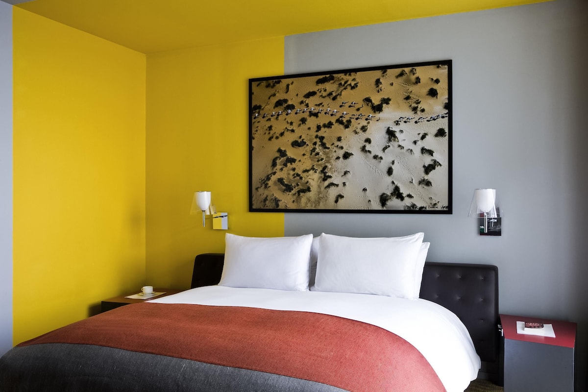 Una habitación de hotel con cama, mesita de noche, lámpara y un cuadro en la pared.