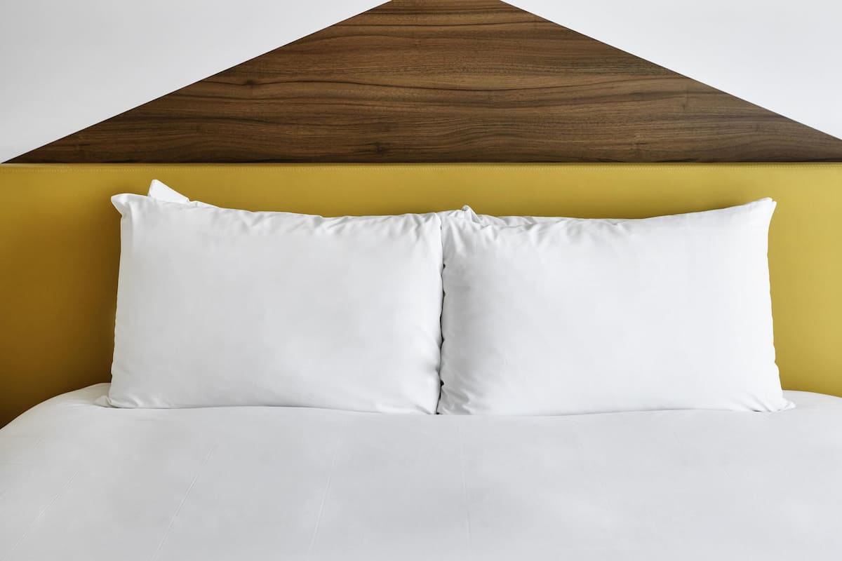 Una cama con ropa de cama blanca y dos almohadas, frente a una pared amarilla con cabecera de madera.
