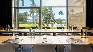 Une salle de conférence dotée de grandes fenêtres donnant sur un espace paysager agrémenté de palmiers et d’une piscine. Des bouteilles d’eau, des verres et des blocs-notes sont à disposition sur les tables.
