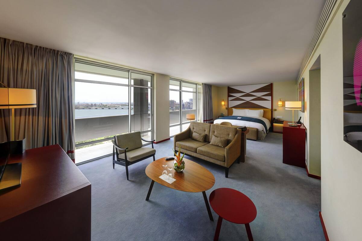 A hotel room with a sofa, a small table, a red chair, a bed, a desk, and large windows offering a city view.
