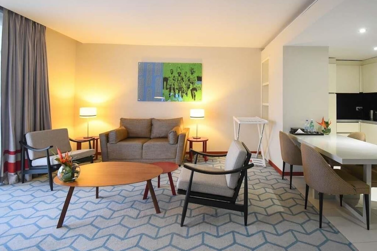 Une chambre d’hôtel comprenant un canapé, deux fauteuils, une table basse, une table à manger avec des chaises, une lampe et un tableau au mur.