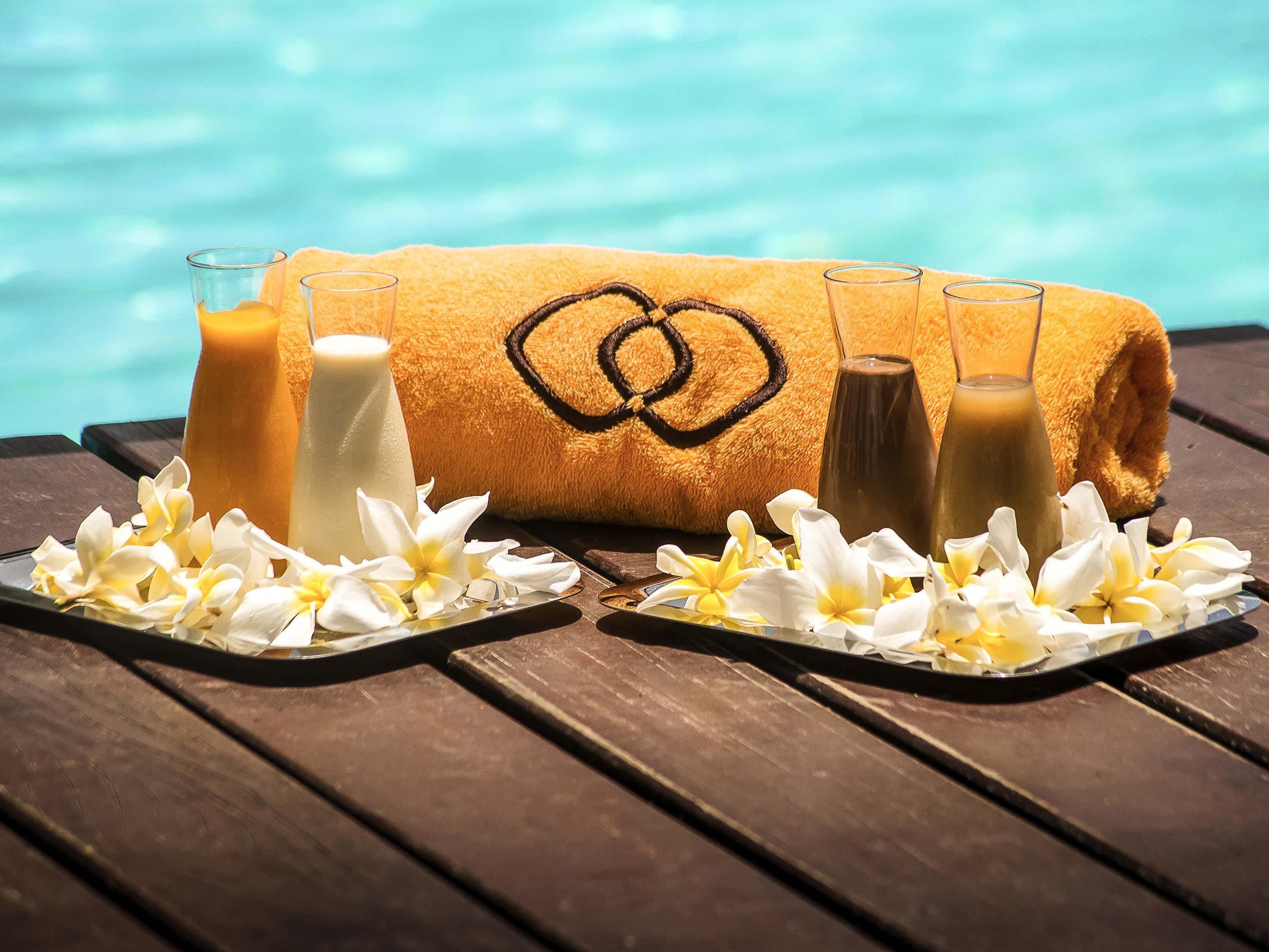 Una toalla con diseño de corazón, dos vasos de bebidas heladas y flores blancas sobre una mesa de madera junto a una piscina.