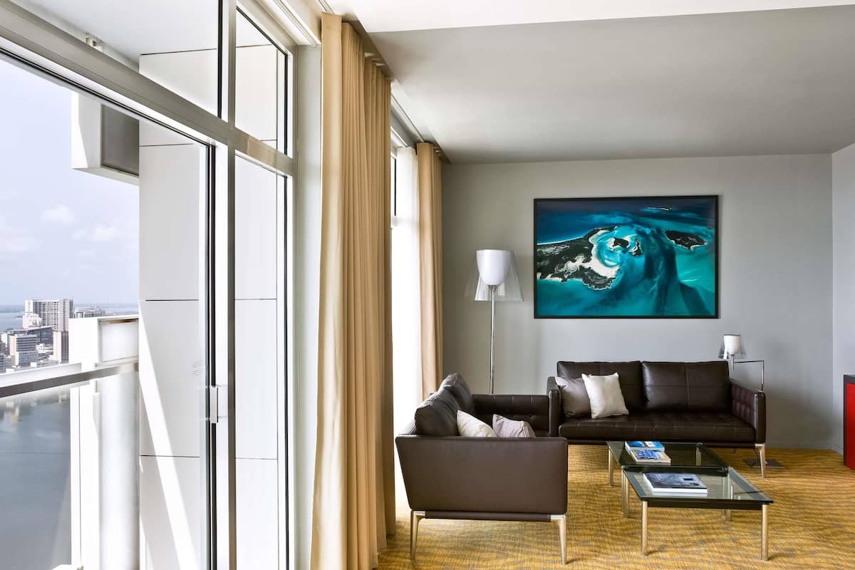 Una habitación de hotel moderna con un ventanal grande, dos sofás, una mesa de centro y un cuadro de una escena oceánica azul.