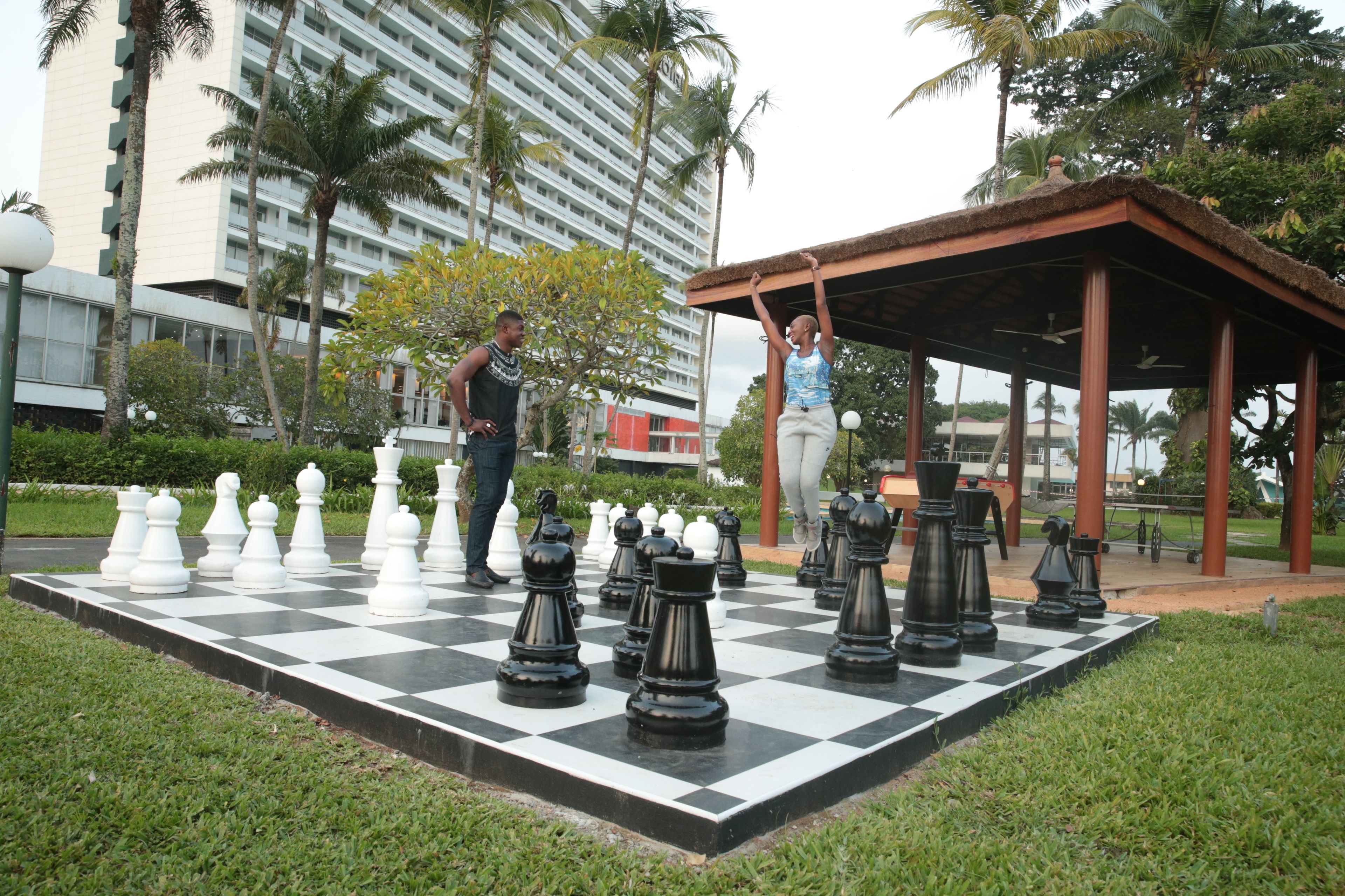 Un jeu d’échecs installé à l’extérieur, avec de grandes pièces d’échecs, entouré de pelouse et de palmiers, et un bâtiment en arrière-plan.