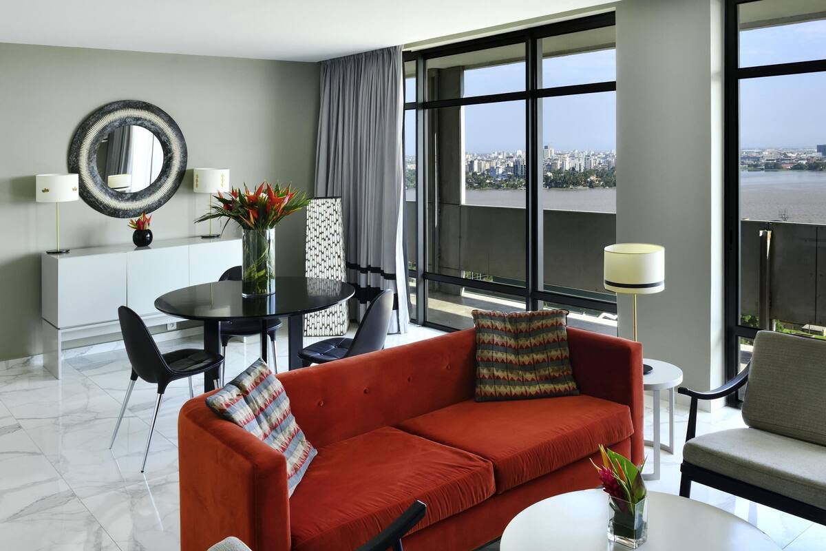 A modern hotel room with a red sofa, a dining table, a large window with a city view, and a white console table with a mirror and lamps.