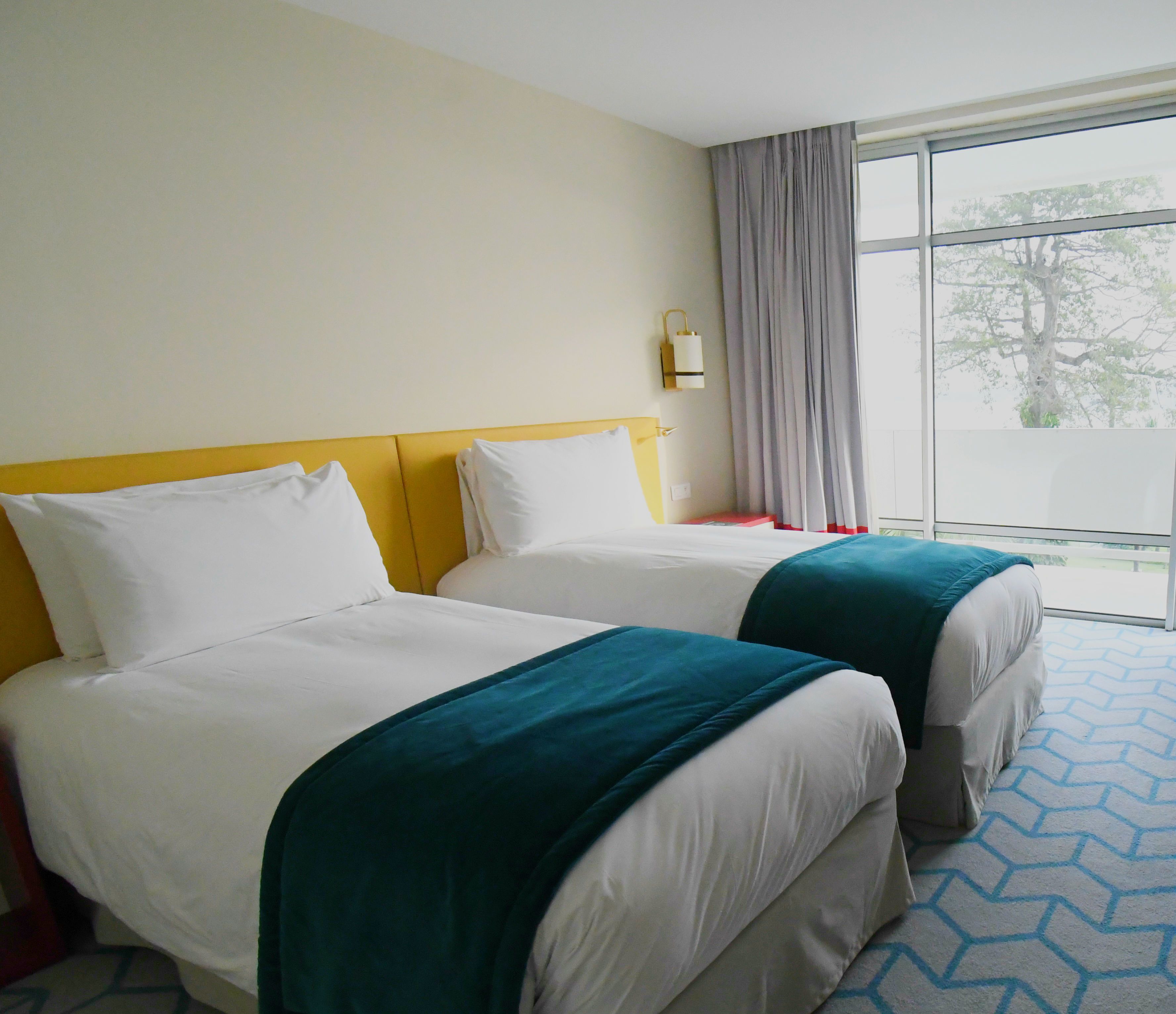 A hotel room with two beds, a large window with curtains, and a view of trees outside.