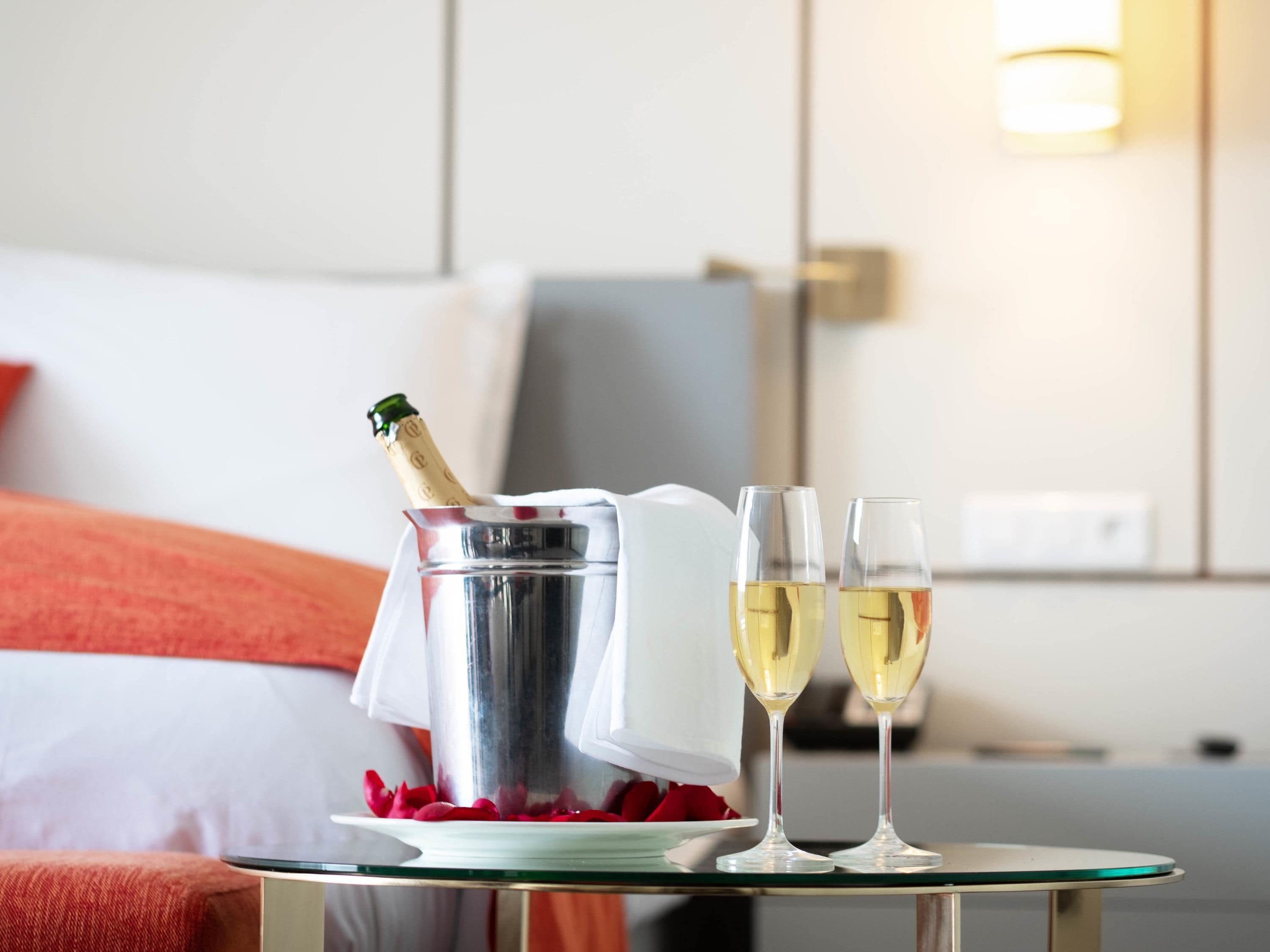 A hotel room with a bed, a bedside table with a champagne bottle and glasses, and a wall-mounted light.