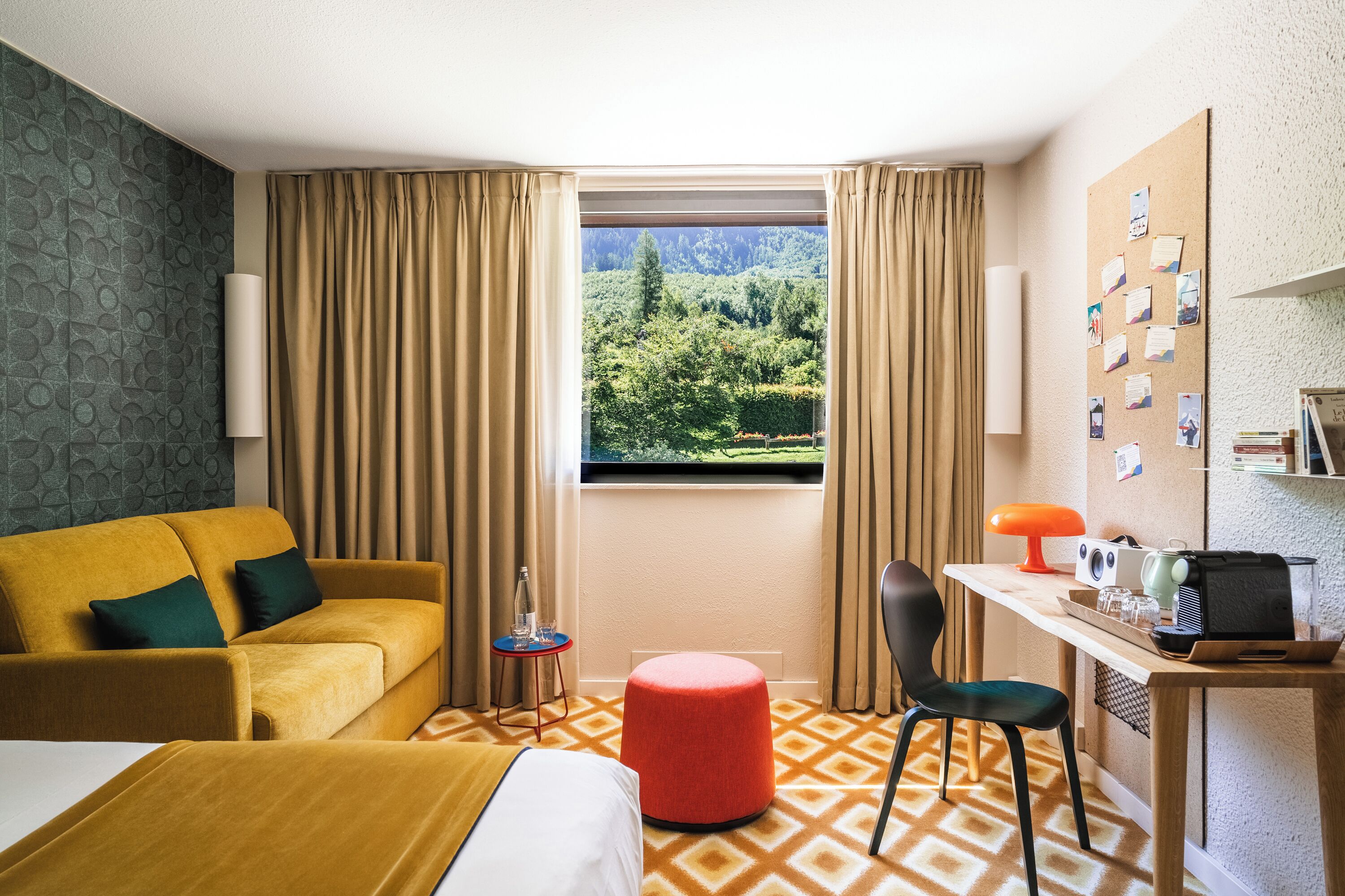 A hotel room with a bed, a sofa, a desk with a lamp, a chair, and a view of a forest through a window.