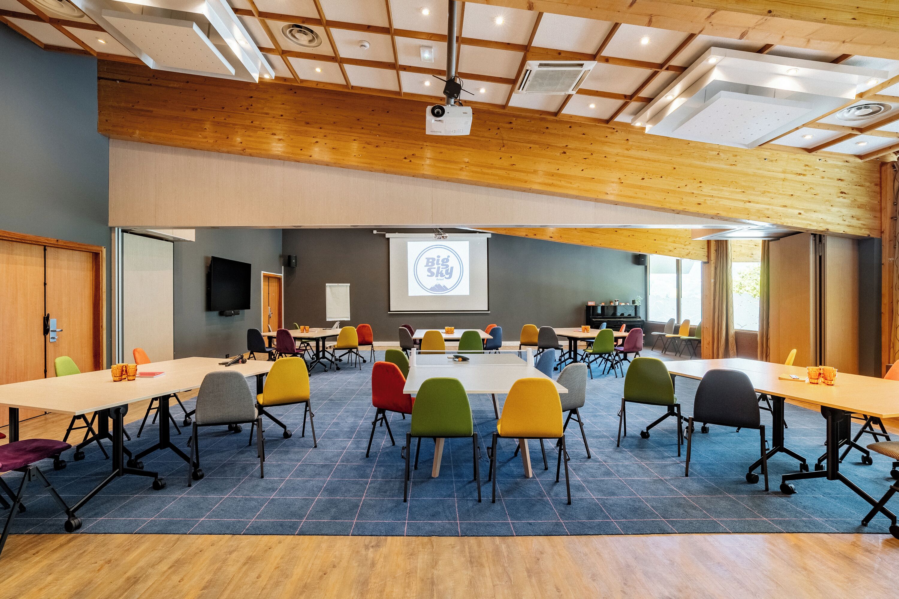 Une salle de conférence dotée d’une grande table, de chaises aux couleurs vives, d’un écran de projection et d’un plafond en bois.