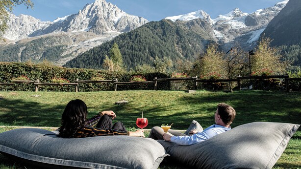 Two people reclining on large gray cushions in a grassy area with a mountain backdrop. -  Big Sky Hotel & Spa (Chamonix-Mont-Blanc)