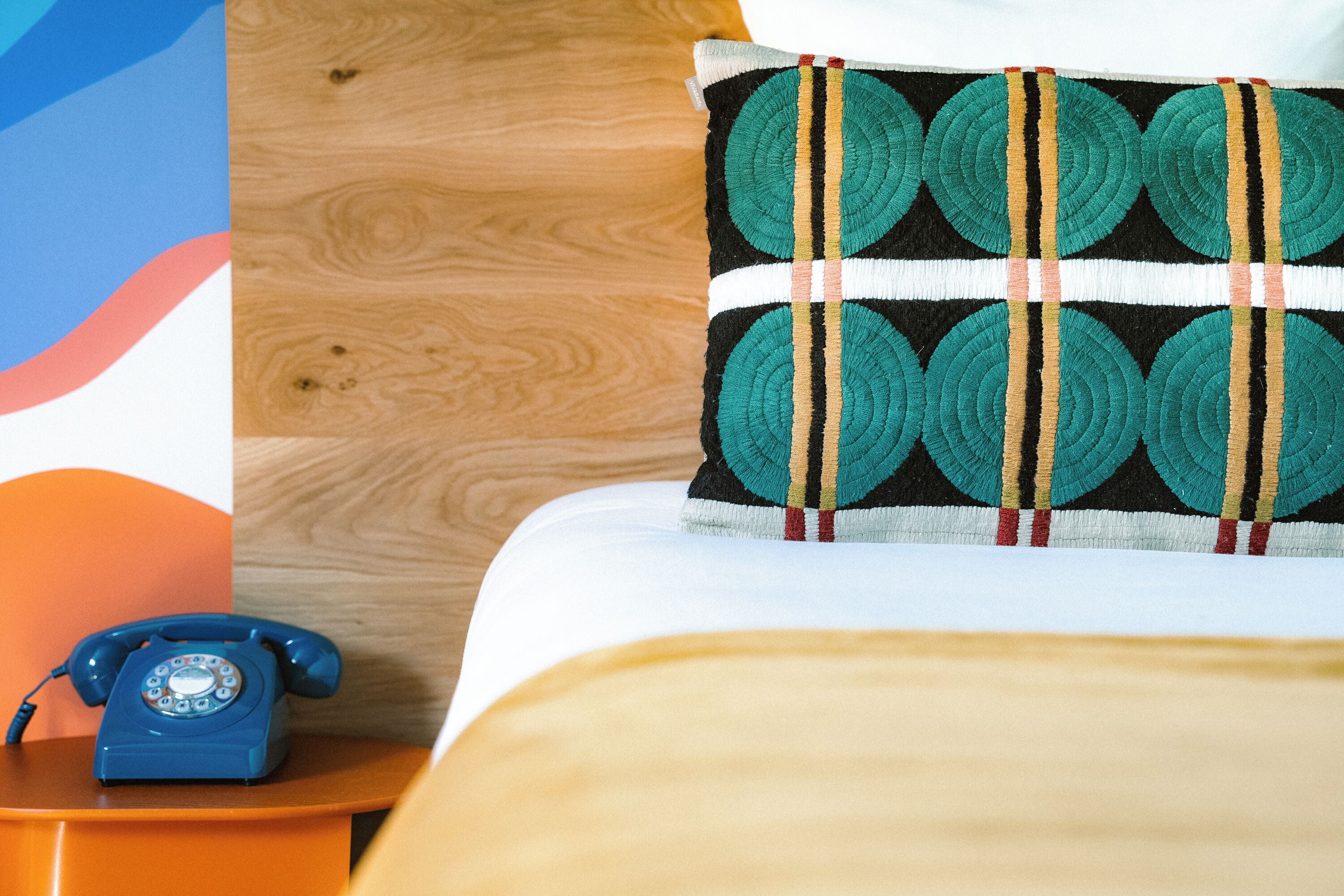 Ein Bett mit einem gemusterten Kopfkissen, einem Holz-Kopfteil und einem blauen, alten Telefon auf einem Nachttisch aus Holz.