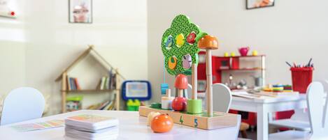 Un área de juegos infantiles con una estructura de juguete de madera, una mesa blanca con material educativo y una estantería al fondo.
