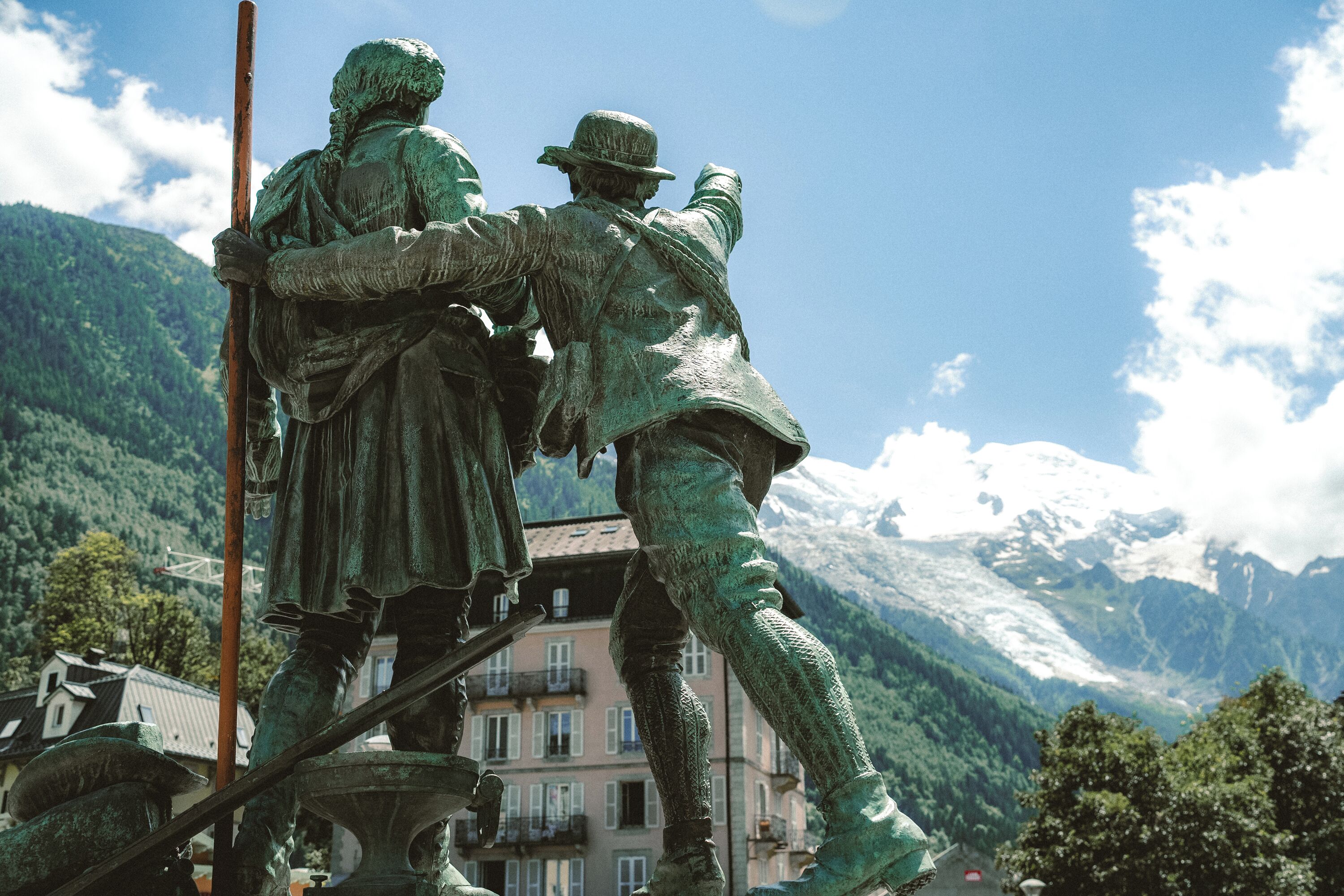 A statue of two figures in historical attire, set against a backdrop of a mountain and a building.