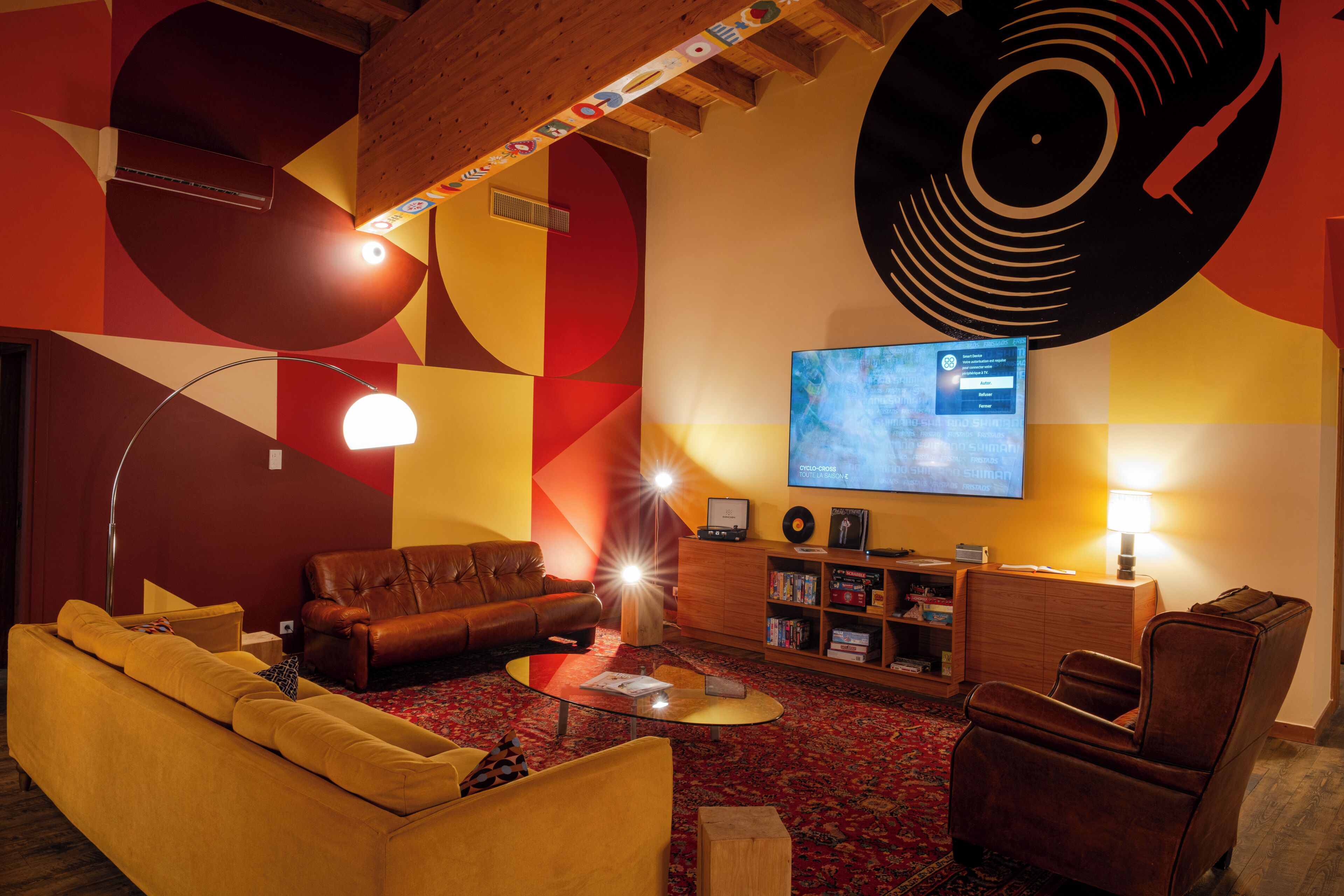 Una sala de estar moderna con un sofá, un sillón y una mesa de centro. Hay un televisor montado en la pared, una estantería y un cuadro abstracto de gran tamaño.