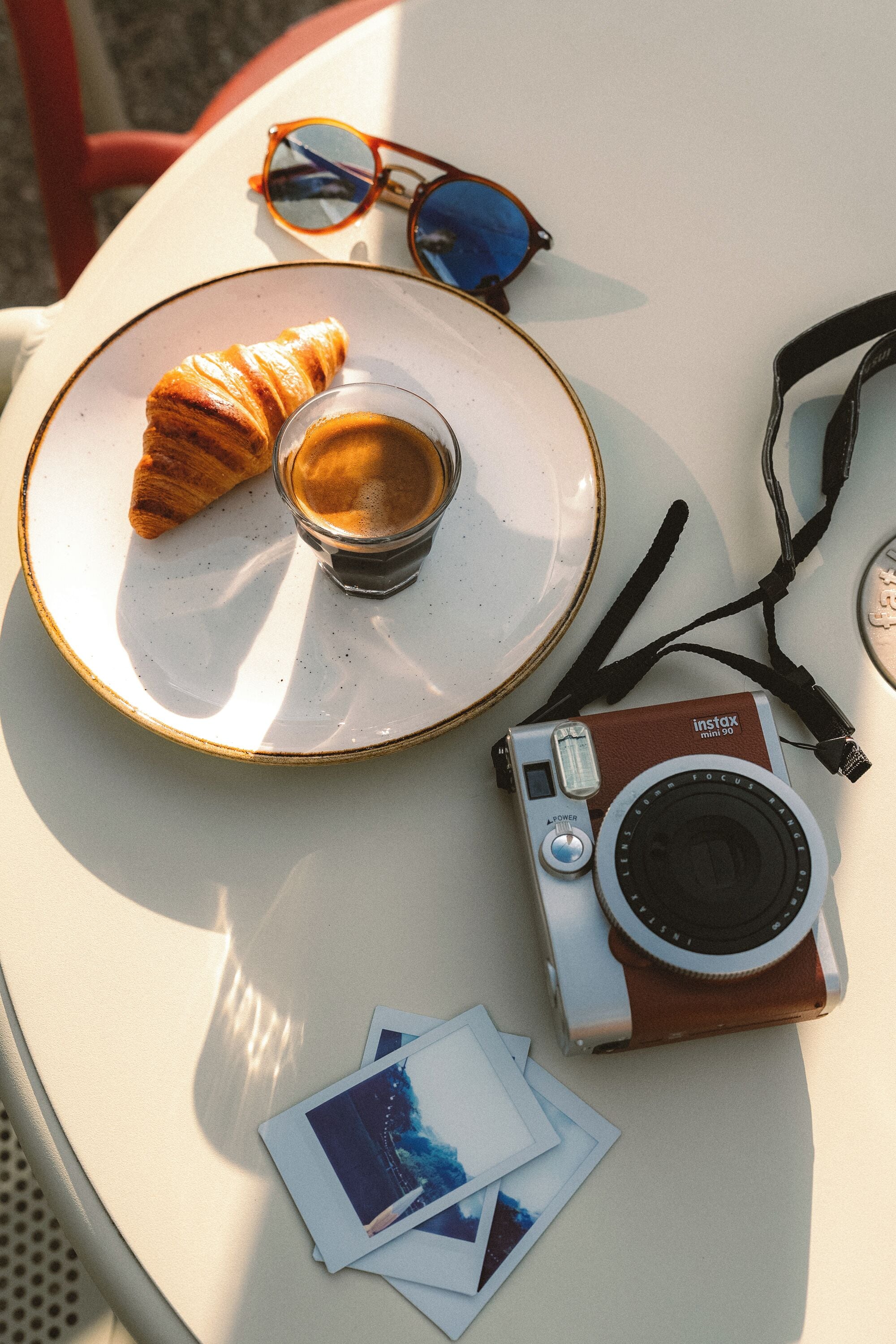 A table with a camera, sunglasses, a croissant, and a cup of coffee.