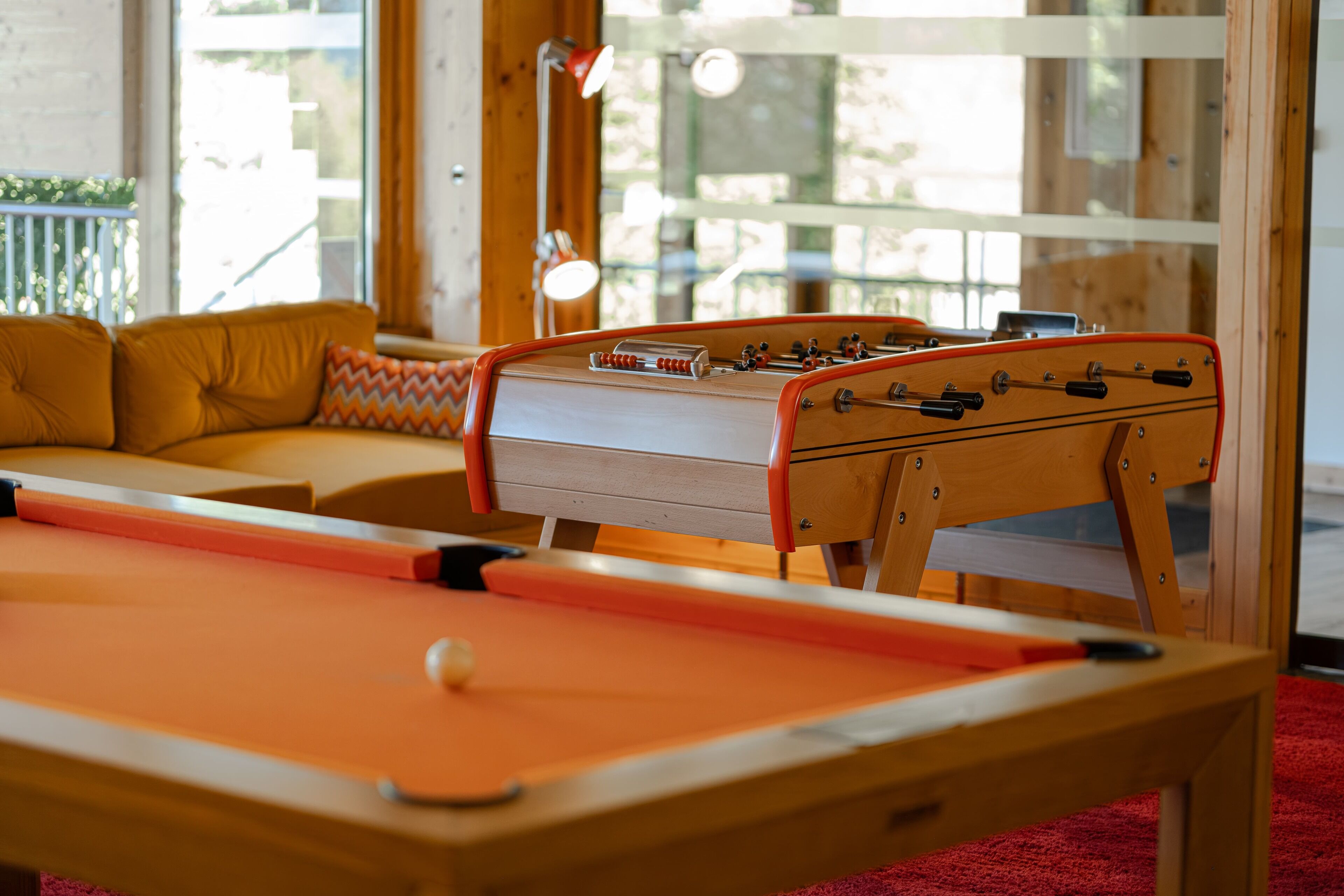 A pool table with a foosball table in a room with wooden furniture and a view of the outdoors.