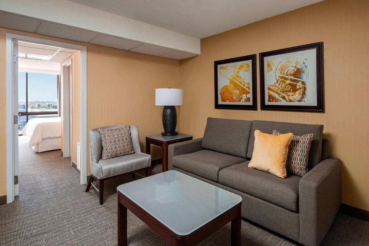 A hotel room with a sofa, a chair, a coffee table, and two framed artworks on the wall.