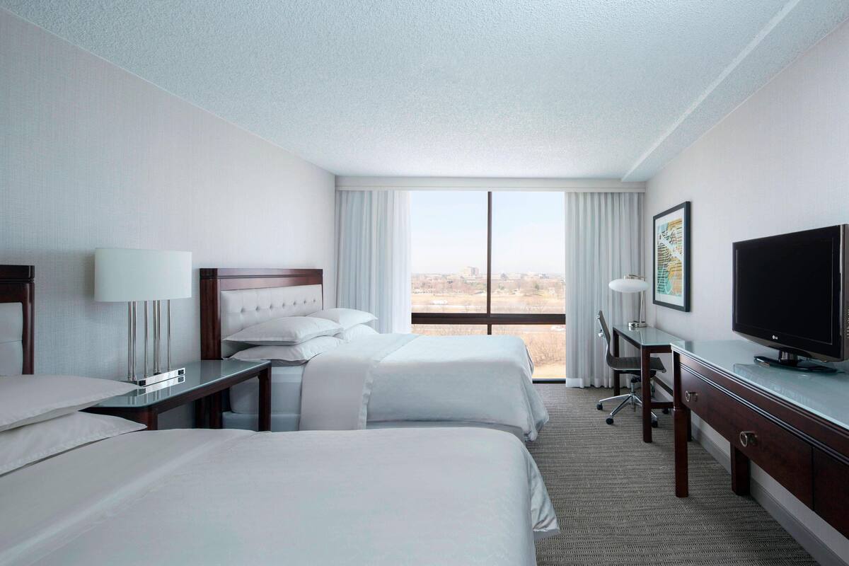 A hotel room with two beds, a desk, a television, and a large window offering a city view.