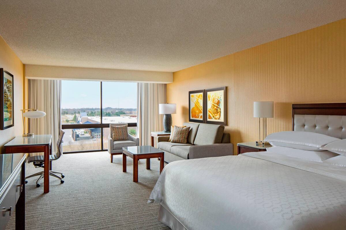 A hotel room with a large bed, a desk, a chair, a sofa, and a view of the city.