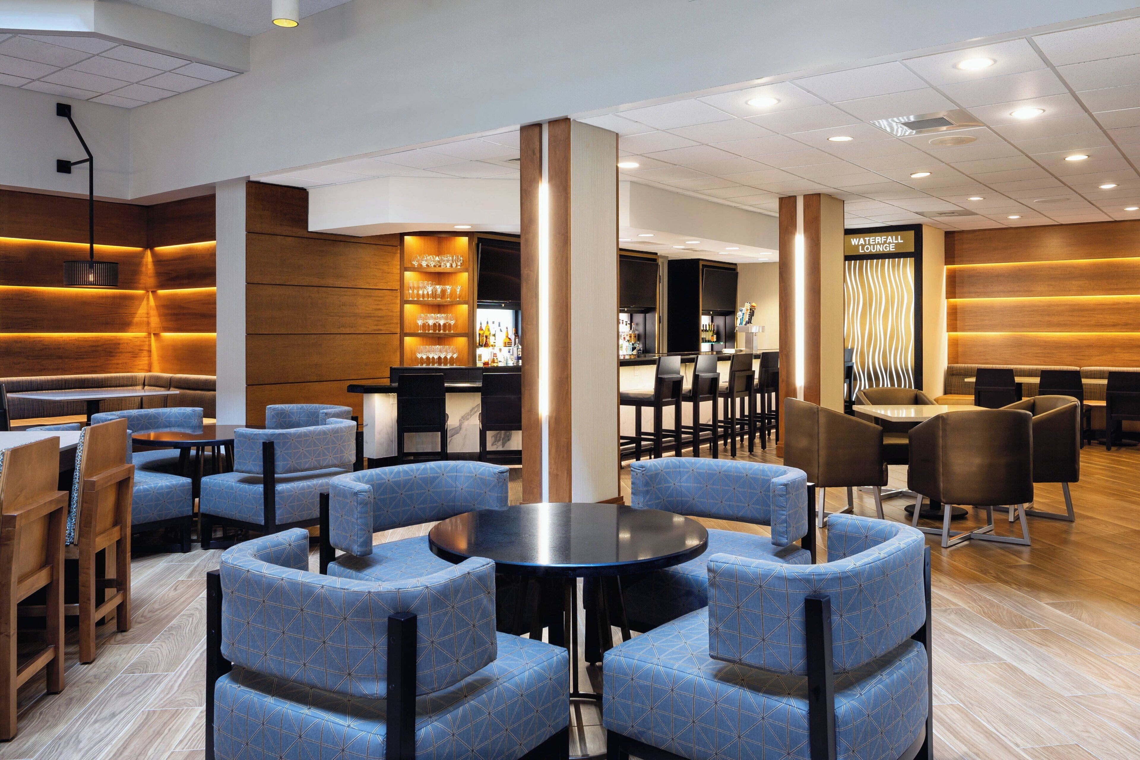 Un salón moderno con sillones de estampado azul, una mesa redonda central y una zona de bar con una cascada.
