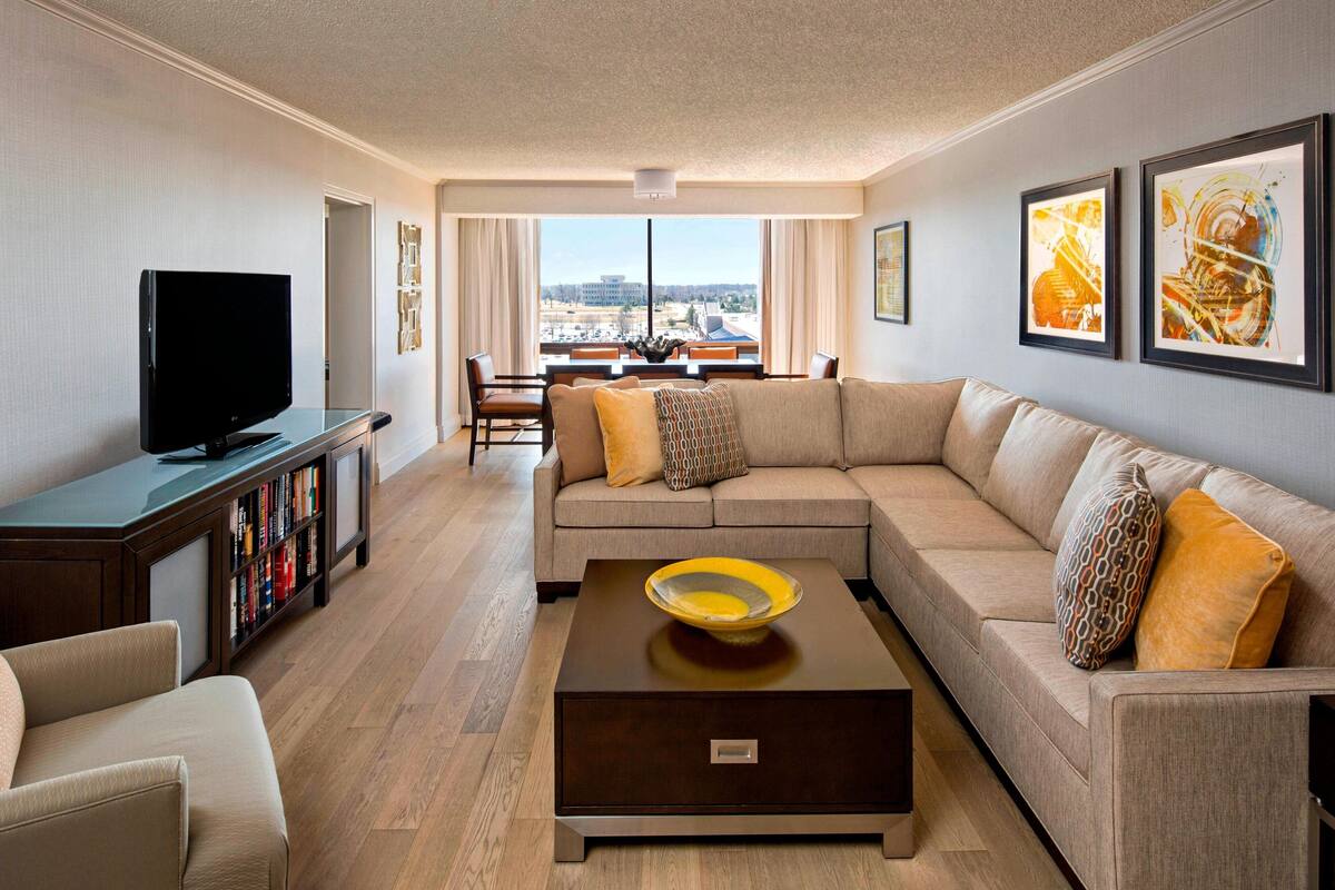 A modern living room with a sofa, coffee table, and TV. There are framed pictures on the wall and a view of the city through large windows.
