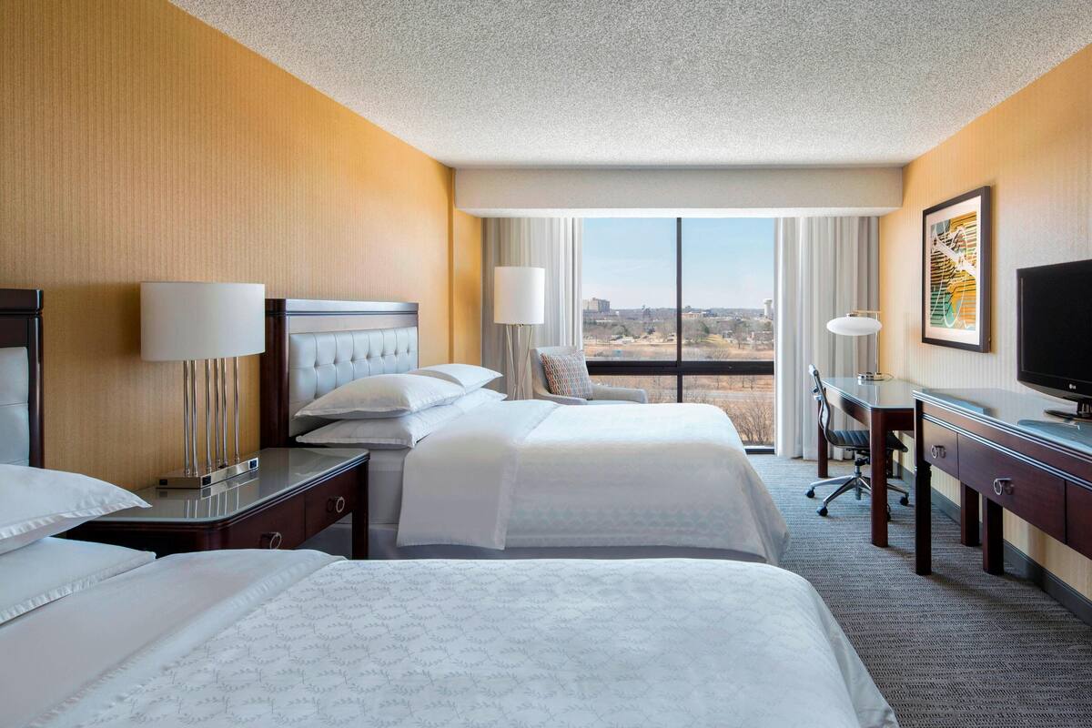 A hotel room with two beds, a desk, a chair, a TV, and a large window offering a city view.