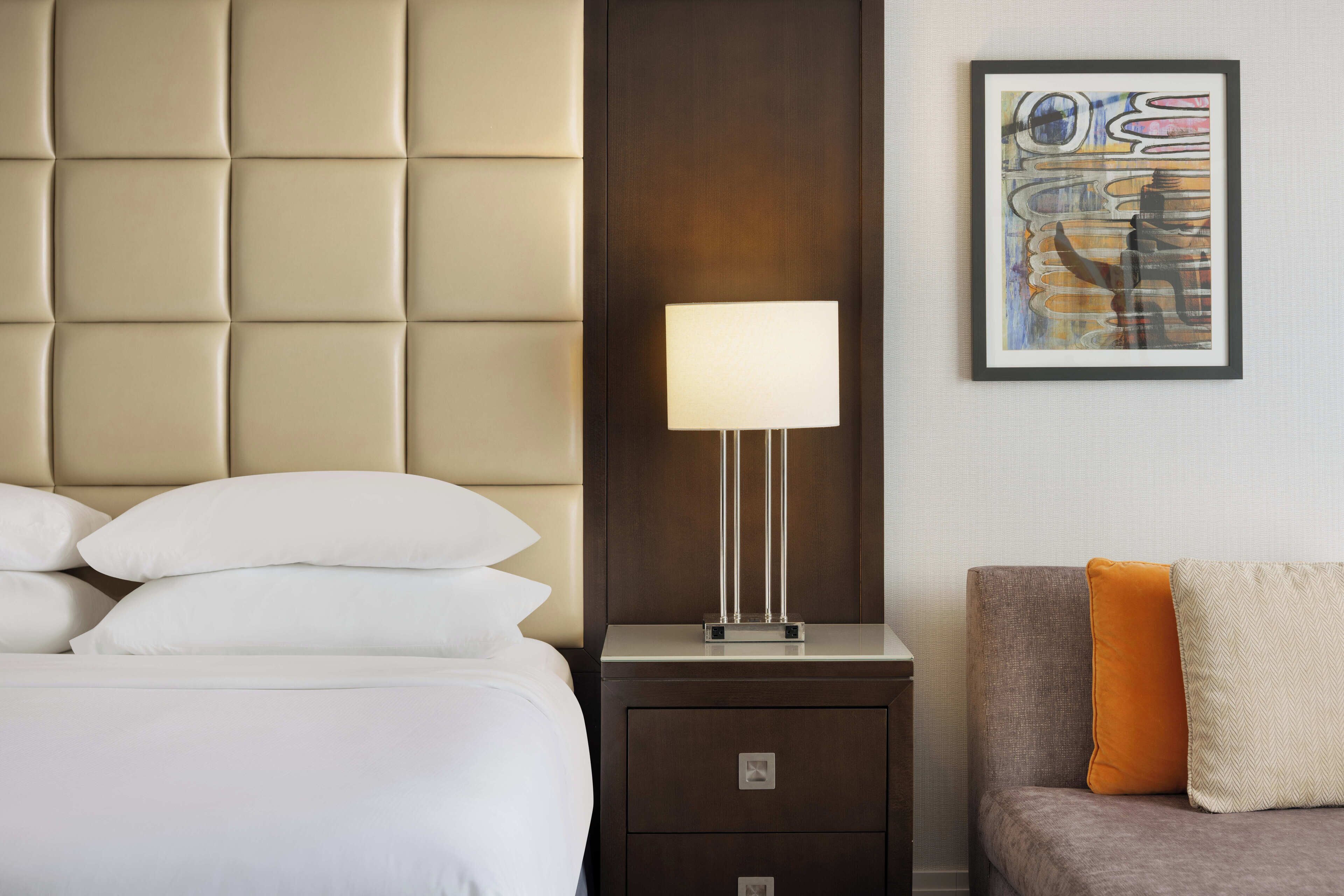 Una camera d'albergo con una testiera imbottita, un comodino con una lampada, un quadro astratto incorniciato e un divano con cuscini.