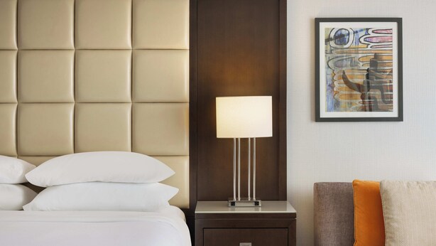 A hotel room with a tufted headboard, a bedside table with a lamp, a framed abstract painting, and a sofa with cushions. - Sheraton West Des Moines Hotel (West Des Moines)